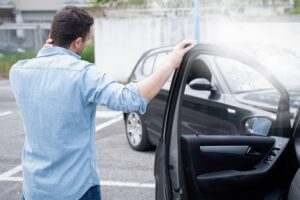 A man stands by an open car door, holding his neck and looking at another car with damage, suggesting a recent accident.