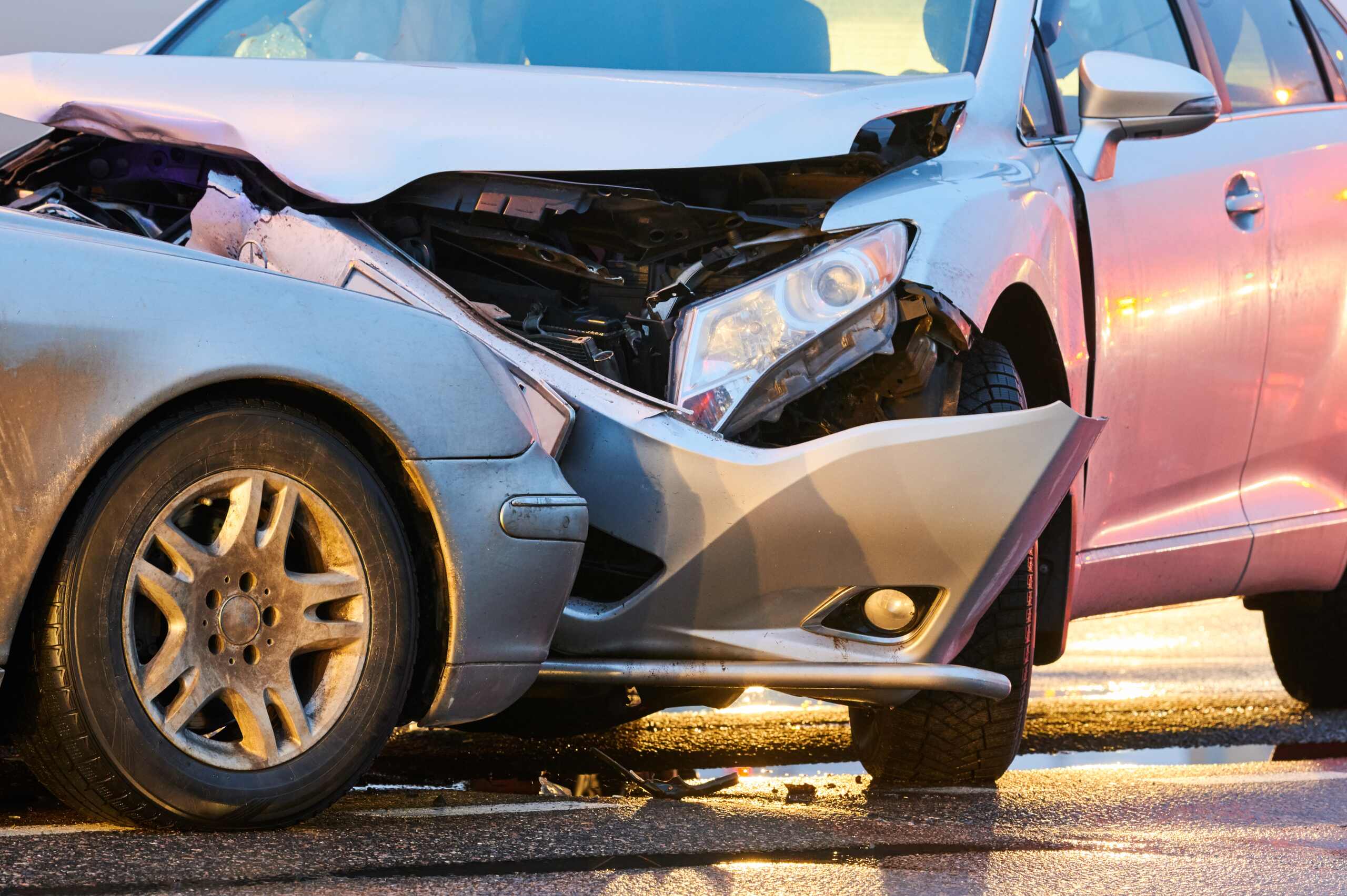 Two silver cars involved in a head-on collision, with significant damage to the front ends of both vehicles, on a wet road.