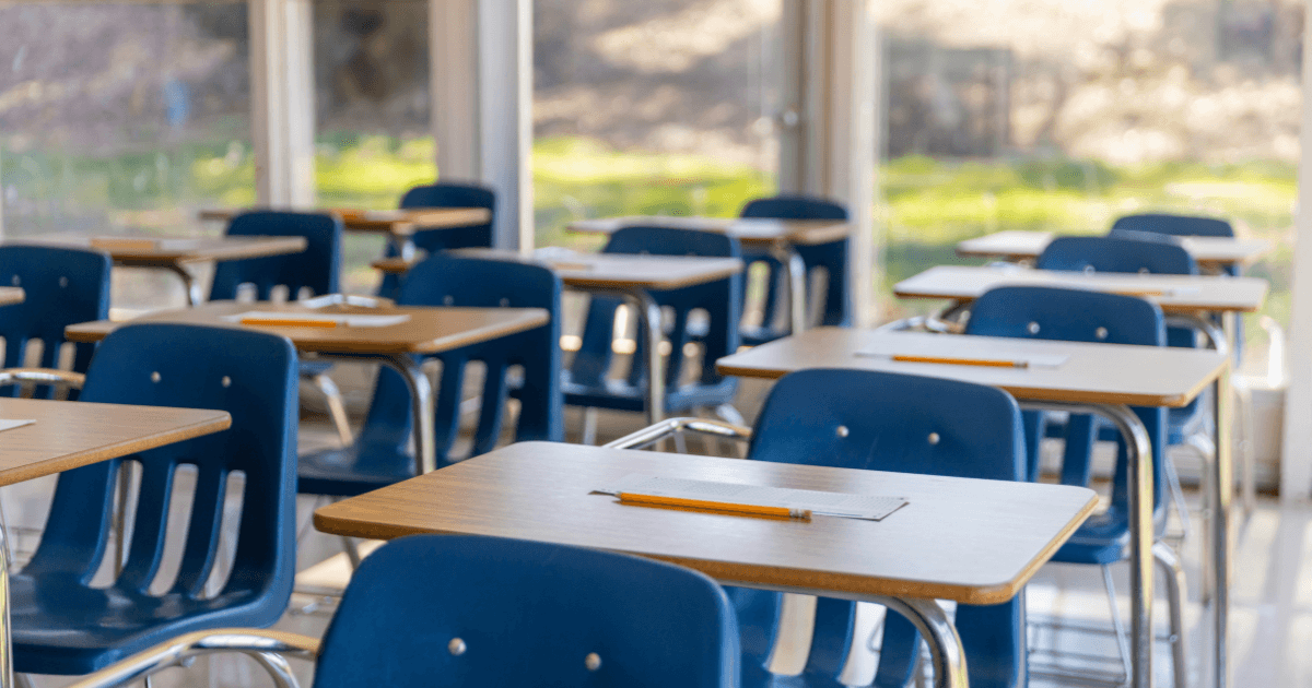 Rows of empty desks with blue chairs in a sunlit classroom, each desk has a sheet of paper and a pencil placed on top.