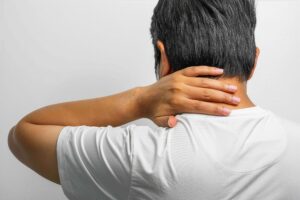 A person wearing a white t-shirt holding the back of their neck, indicating discomfort or pain.