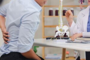 A doctor points to a model of a human spine while consulting with a patient who is holding his lower back.
